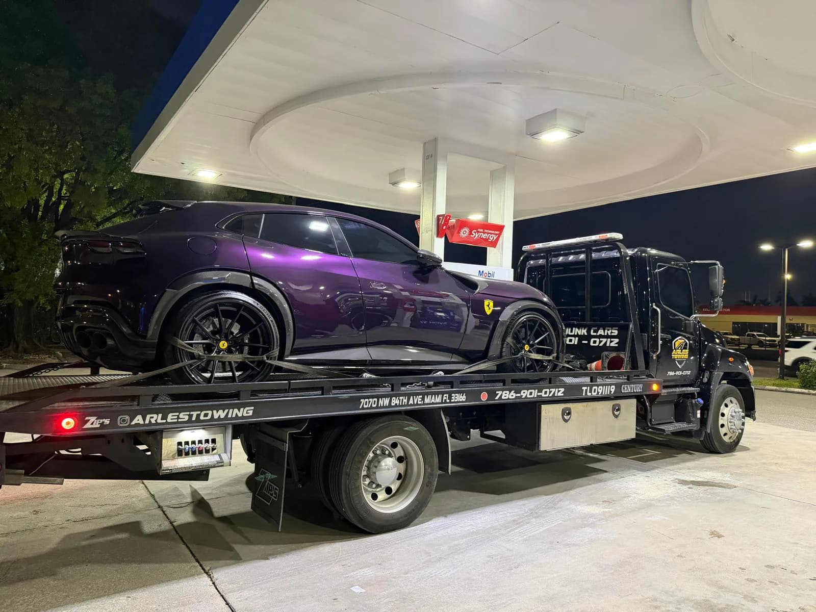 Arles Towing black Hino flatbed carrying a purple Ferrari Purosangue at a Miami gas station at night